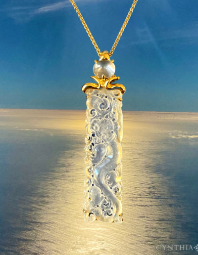 Pendant featuring a hand-carved Icy Jade rectangle crowned with a blue sheen Moonstone, set in 18 karat yellow gold suspended over a calm sea with golden light from above